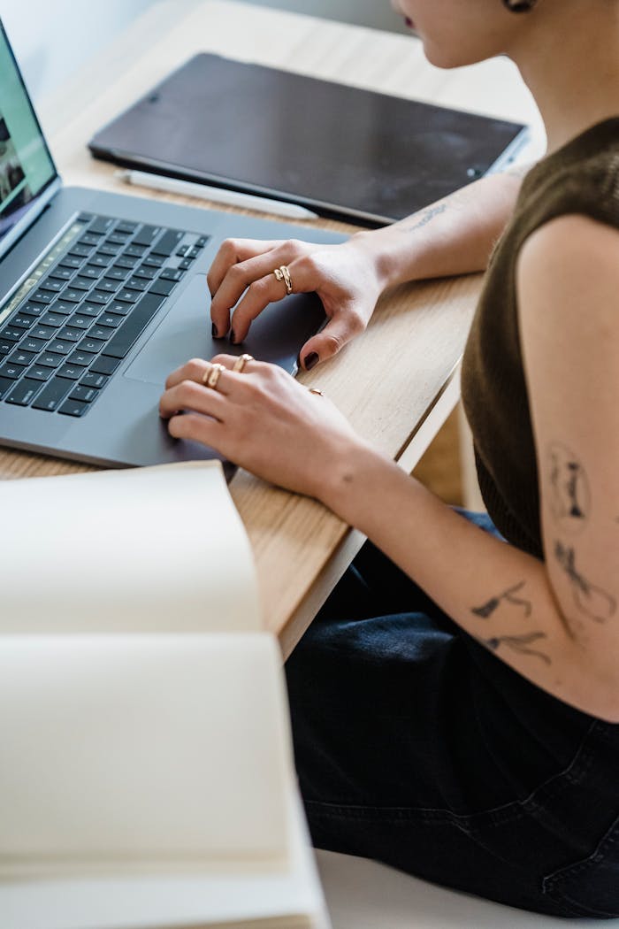 our-experience Side view high angle of crop faceless woman in casual clothes with tattoos sitting on chair at table while working remotely on netbook near sketchbook and tablet in light apartment